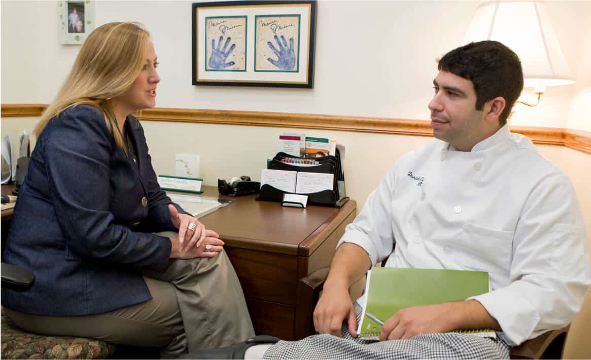 A woman in a business suit and a man in a chef's uniform sit in an office, engaged in conversation about student life. The man holds a green folder on his lap. Behind them, on the wall, are framed children's handprints, and a lamp is on the desk.