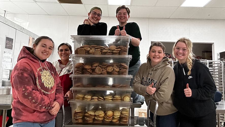 A group of six people, wearing casual clothes and aprons, smile and give thumbs up in a kitchen. They stand in front of a large rack of baking trays inside an industrial oven. The setting appears to be a commercial kitchen or bakery.