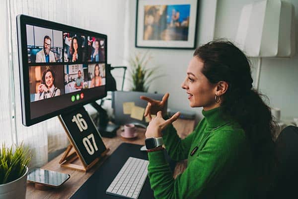 A young woman in a green sweater is sitting at her desk and engaging in a video conference with her online culinary school. The clock on her desk shows 16:07, and the desk is decorated with plants, office supplies, and a lamp.