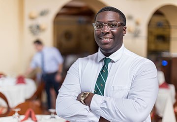 A confident man stands with arms crossed, wearing a white shirt and a green tie, smiling in a well-lit restaurant. A blurred figure is seen in the background, setting up a table with red napkins. The ambiance suggests a formal dining environment.