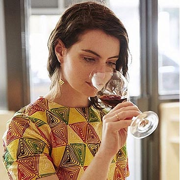 Andrea Morris, a CIA alumna with dark hair, is indoors wearing a colorful patterned shirt. She's concentrating on the aroma of a glass of red wine, with the background softly blurred.