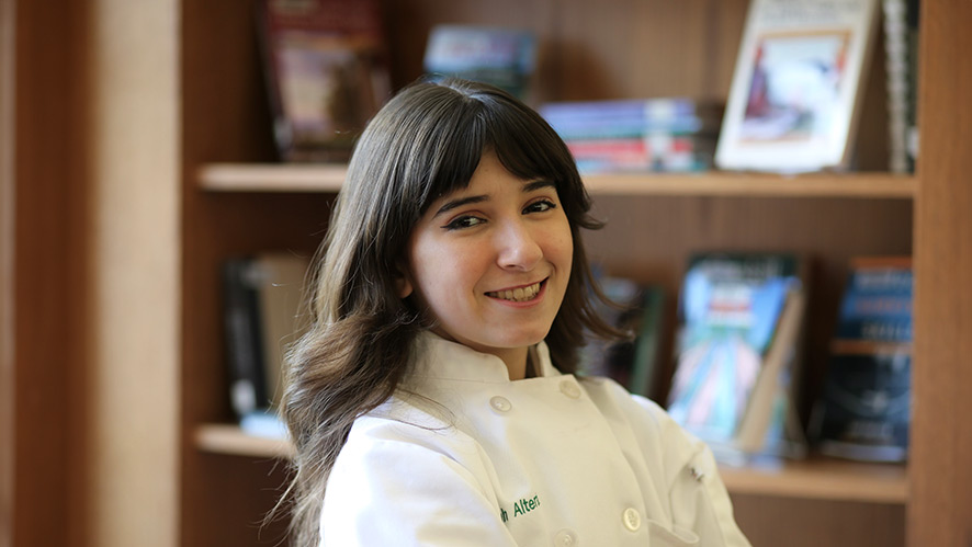 A smiling Deborah Alterio in her chef whites with a bookshelf behind her.