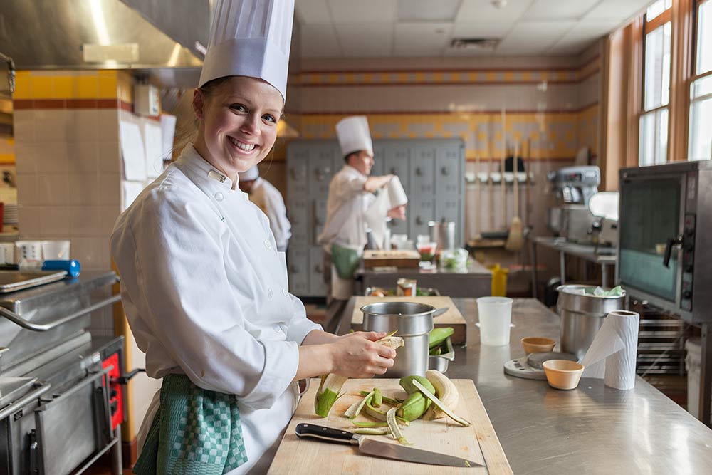 Image of a smiling CIA student peeling plantains at a cutting board standing behind a counter in a kitchen.